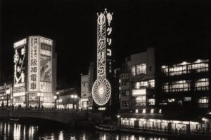 Black-and-white historical reconstruction of the original Glico Running Man sign overlooking Osaka’s Dotonbori canal in the 1930s, surrounded by prewar buildings and advertisements.